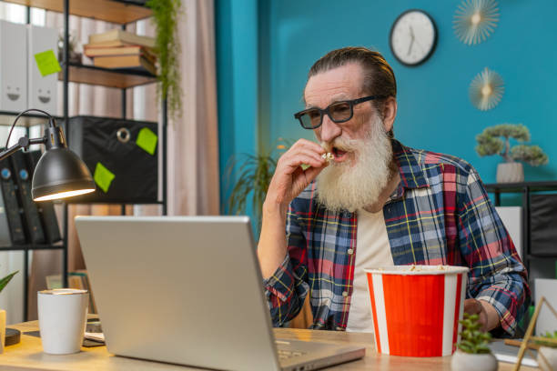 Senior man freelancer taking break from work wearing 3D glasses eating popcorn watching movie film