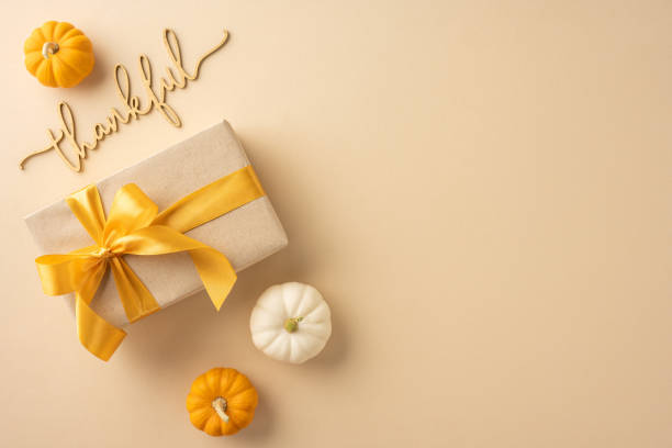 A Thanksgiving themed gift box with a yellow ribbon, surrounded by small pumpkins, and a thankful message on a beige background