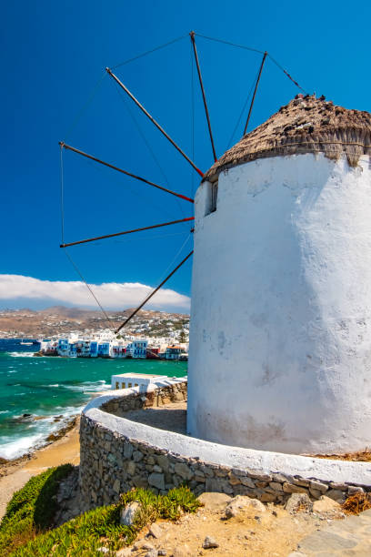 An old traditional whitewashed windmill with a thatched roof on the Greek Island of Mykonos, Greece. Set against a blue cloudy sky and the Aegean Sea.