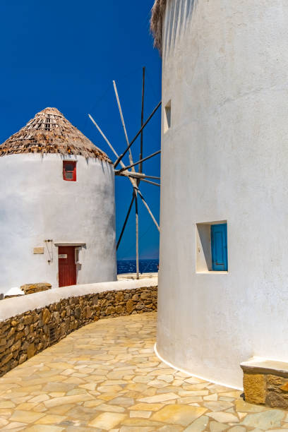 An old traditional whitewashed windmill with a red door and a thatched roof on the Greek Island of Mykonos, Greece. Set against a blue cloudy sky and the Aegean Sea.