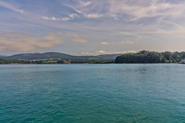 A serene view of a calm lake surrounded by lush green hills under a partly cloudy sky. The water reflects the clouds and greenery, creating a peaceful landscape.