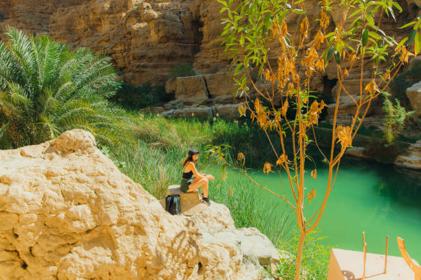 Middle Eastern Woman Contemplating in Scenic Wadi Shab Canyon and Majestic Landscapes of Oman Nature Side view of female traveller with backpack in black sport clothing sitting with picturesque viewpoint of Wadi Ash Shab canyon and emerald green river, admiring desert Middle Eastern landscapes in Oman during sunny day wadi shab stock pictures, royalty-free photos & images