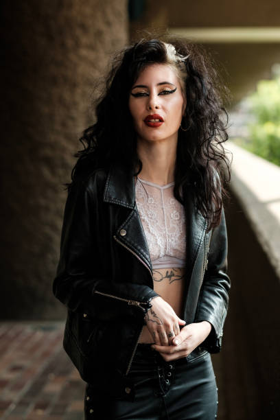 Female rocker posing in an urban environment. She is wearing black leather outfit and white top. Her hands are together and she is looking at camera.