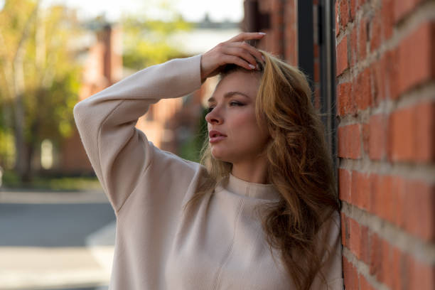 Close-up side view portrait of beautiful serene long haired caucasian blond woman in blank white sweater standing outdoor on city street by brick building in a sunny autumn morning. Fashion theme.