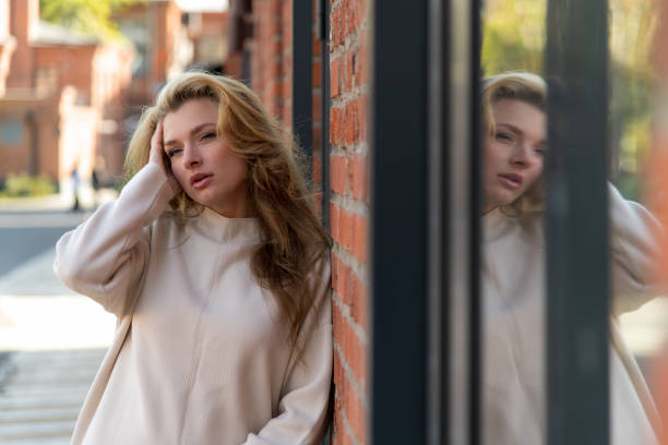 Close-up portrait of beautiful serene long haired blond woman in white sweater standing outdoor on city street by red brick loft building wall and reflects in window in autumn day. Fashion theme.