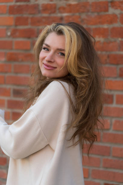 Attractive young blond-red haired woman standing in front of white brick wall hands behind her head looking towards the camera.