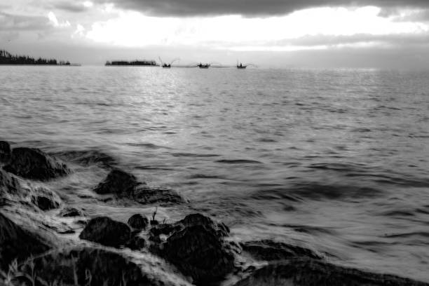 A black and white photo of several fishing boats on a calm body of water, silhouetted against a dramatic sunset. The image evokes a sense of peace and tranquility, as the day comes to a close.
