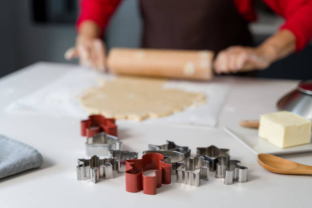 Cookie cutters on kitchen table with woman rolling dough for christmas cookies in background
