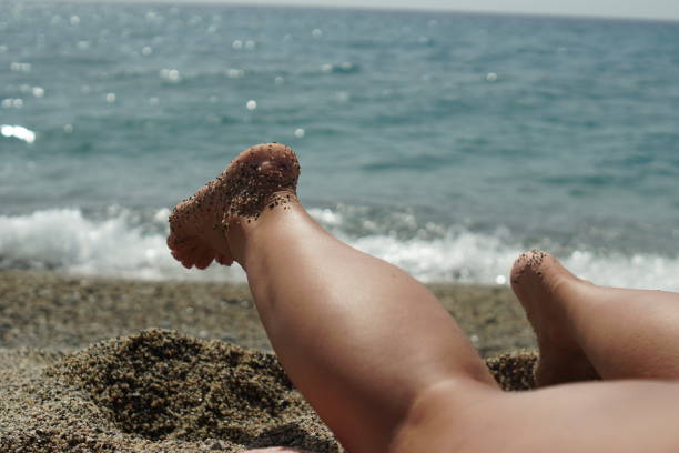 Women's feet immersed in soft sand. Relaxation and serenity on the beach on a warm summer day. A girl lies on the sand by the sea, swinging her legs.