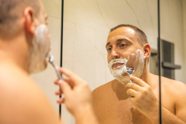 istock Man with shaving foam on his face standing in bathroom, looking at mirror, shaving his face beard. 2178246748