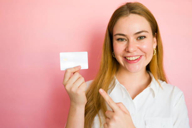 istock Smiling young woman pointing at a credit card while holding it on a pink background 2178272476