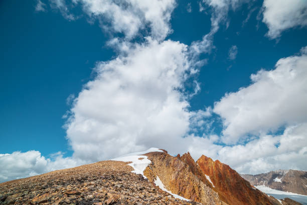 istock Snow-capped mountain ridge with cornice and red sharp rocks on top in sunlight. Snow cap and shiny pointy peak of gold color. Snow dome and colorful rocky peaked top. Sunny cloudy high mountains view. 2178273434