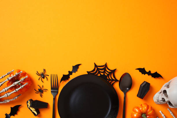 istock Halloween table with black empty plate, cutlery, spooky decorations, bats, pumpkins, skeleton arms, skull on orange background. Flat lay, top view. 2178276391