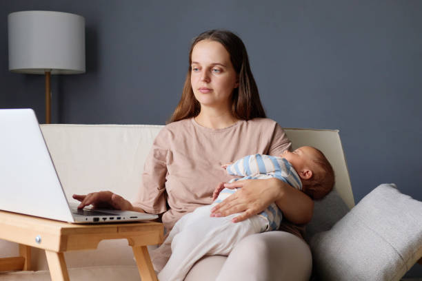 istock Focused woman sitting on couch typing on her laptop while balancing business tasks and motherhood with her cute baby in hands working remotely during maternity leave in her cozy home workspace 2178299043