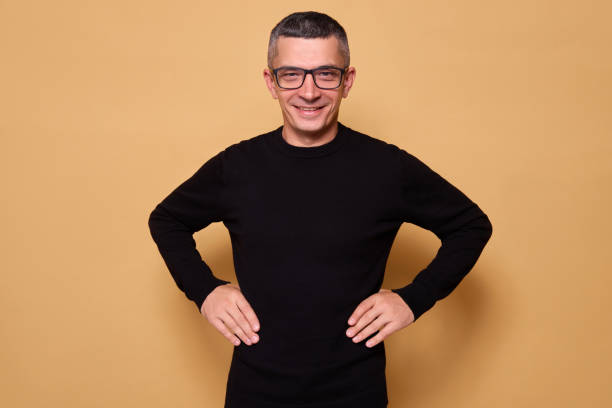 istock Confident man wearing black raglan with glasses standing with hands on hips smiling with toothy grin showing joy and satisfaction radiating friendly and optimistic vibe isolated over beige background 2178299897