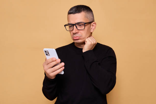 istock Upset man in glasses and jumper looking sad while using his smartphone to read messages and connect online demonstrating feeling of sorrow isolated over beige background 2178299914