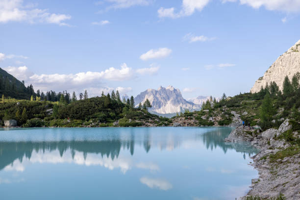 A peaceful turquoise lake with a forested shoreline, reflecting the surrounding trees and distant rocky peaks.