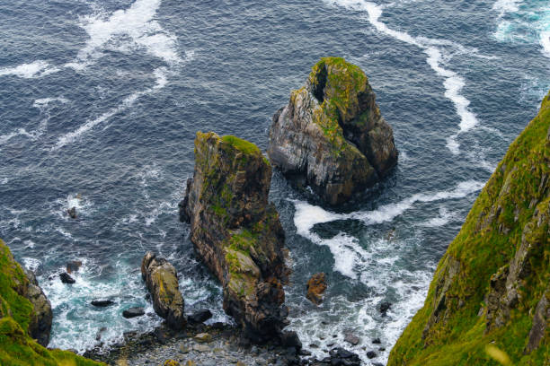Majestic rock formations emerging from the sea, covered in lush green moss, surrounded by foamy ocean waves.