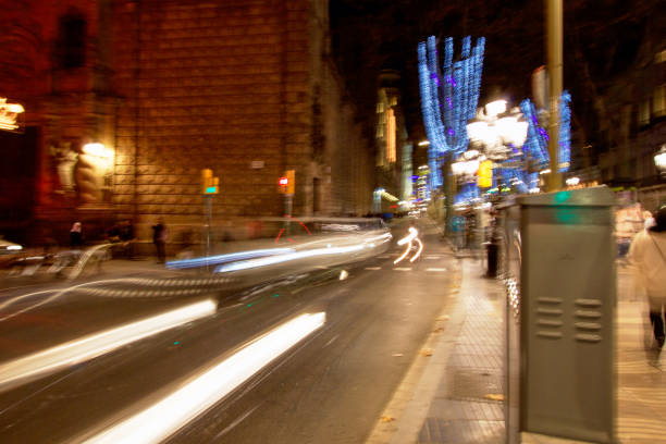 This dynamic photograph captures the energy of a busy Barcelona street at night, with the motion of passing cars transformed into bright streaks of light. The surrounding buildings, adorned with illuminated trees and festive decorations, add a sense of celebration to the scene. The soft blur of light trails creates a feeling of movement and speed, while the stationary elements—street lamps, traffic lights, and the architecture—ground the image in place. The contrast between the bright lights and the darker surroundings evokes the vibrant pulse of the city as it comes to life after dark.