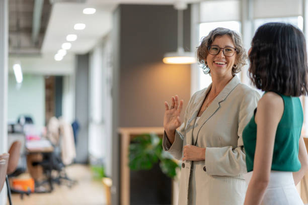 istock Experienced business developer standing with a young female employee, sharing a friendly conversation with a bright smile in a modern office. Training and task handover take place in a supportive, collaborative environment focused on teamwork, success. 2178329608