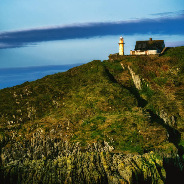 A white lighthouse stands on a rocky cliff, next to a small, white, single-story house with a grey roof. The lighthouse is tall and slender, with a white cylindrical body and a black lantern.