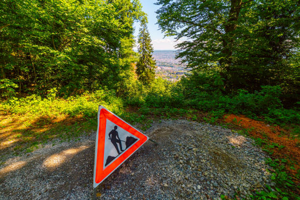 A triangular road work sign stands on a gravel path in a tranquil forest, overlooking a distant city. The bright red sign contrasts with the lush greenery.