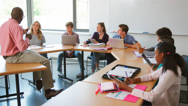 istock Male High School Tutor Sitting On Desk And Asking Students With Laptops And Digital Tablets Question 2178336991
