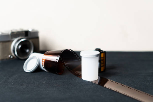 A vintage camera with a roll of film and a white film canister on a dark surface. The camera is slightly out of focus in the background, while the film and canister are prominently displayed