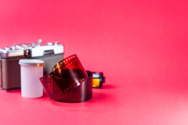 A vintage camera with a roll of film and a film canister on a bright pink background. The camera is metallic with a classic design, and the film is partially unrolled, showcasing its perforated edges.