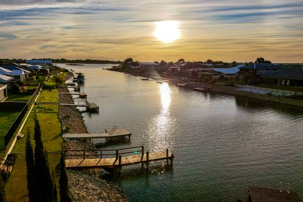 This serene waterfront scene captures the beauty of residential living along a peaceful canal. The sun sets over the horizon, casting a golden reflection across the calm water. Modern houses with private docks line the water’s edge, offering scenic views and direct access to the canal. The vibrant sky, with streaks of clouds, enhances the peaceful ambiance, creating a perfect moment for relaxation by the water. The image portrays a blend of nature and modern life, showcasing a tranquil lifestyle by the waterfront.