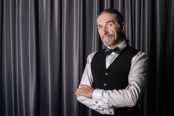 Portrait of an older man with a beard and tied-back hair, wearing formal attire with a bow tie and vest, posing confidently by curtains