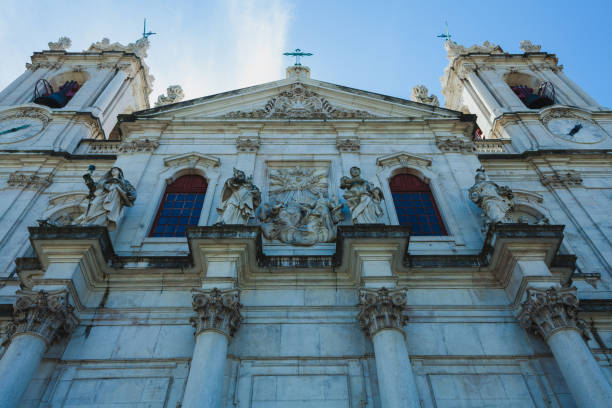 the grand façade of a historic church or cathedral, featuring intricate architectural elements and several sculptures adorning the structure. The stone building is characterized by tall columns with Corinthian capitals and an array of statues placed in niches or on ledges. Above, a detailed relief in the center depicts a religious scene, possibly of heavenly figures. The buildings towering spires and arched windows, framed by ornate carvings, contribute to the imposing and majestic appearance of the structure.