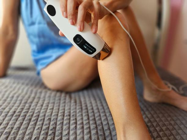 Young woman in bathrobe epilating her legs with modern photoepilator Young woman in bathrobe epilating her legs with modern photoepilator on sofa at home Best Laser Hair Removal Products stock pictures, royalty-free photos & images
