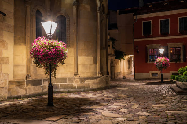 Illuminated cobblestone street with blooming flower baskets on lampposts outside the Armenian Cathedral in Lviv. Historic architecture and warm lighting create a romantic atmosphere