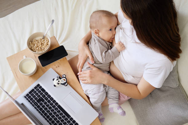 istock Young mom embracing infant and browsing data on laptop sitting on bed and working on remote project at home while breastfeeding her infant son or daughter 2178384455