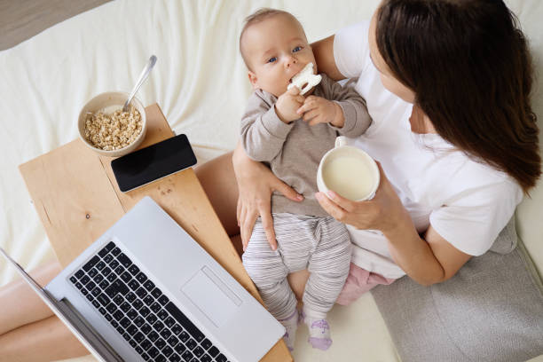 istock Mother sitting in bedroom working on computer while or doing online shopping for children and bonding with newborn playing peacefully in her hands enjoying cup of coffee 2178384458
