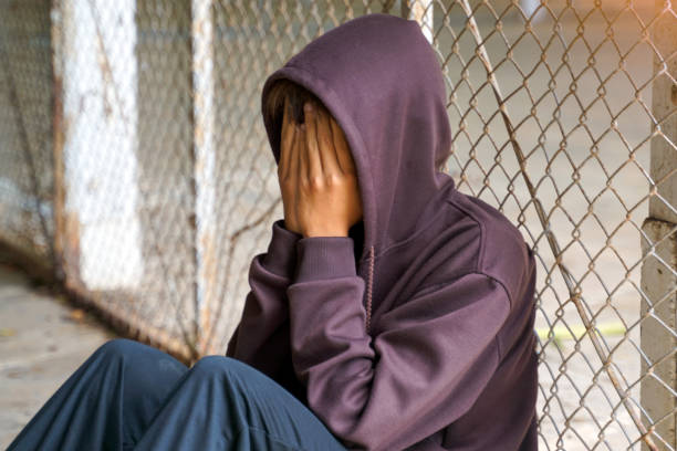 Young man wearing a hood over his head. He covered his face with his hands and sat alone against the mesh fence.