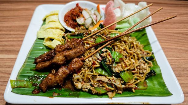 istock Stir-fried noodles served with grilled chicken skewers, omelette slices, and assorted vegetables on a banana leaf-lined plate with sambal and crackers on the side, presented on a wooden table 2178387965