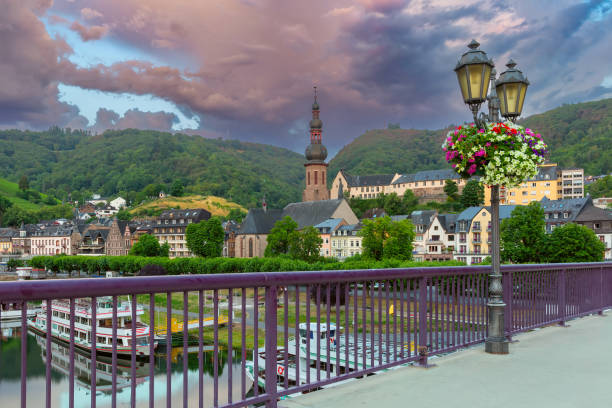 View of Cochem bridge with lamp post, church, and colorful buildings under dramatic sky, Germany