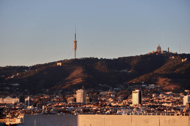 This image presents a panoramic view of Barcelona with the iconic Collserola Tower standing tall on the hill. The tower is a prominent part of the city’s skyline, with Tibidabo amusement park and its cathedral, the Temple Expiatori del Sagrat Cor, visible in the background on the right. The city unfolds below, showcasing the densely packed buildings of Barcelona with sunlight softly illuminating the scene in the late afternoon or early morning. The contrast between the urban landscape and the natural hills in the background creates a striking composition, highlighting the balance between nature and architecture in the city.