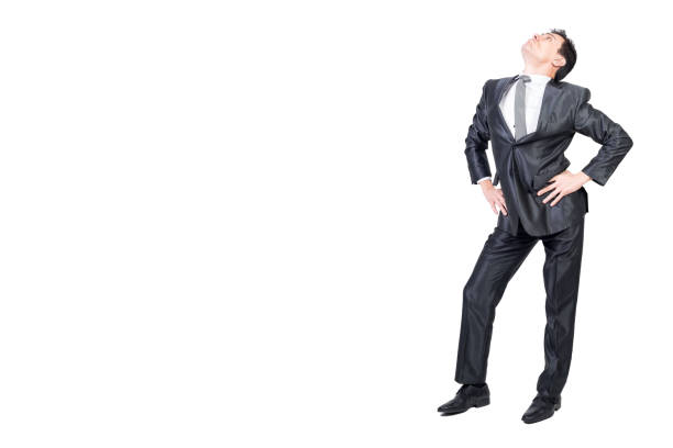 Full body of young arrogant male manager in classy suit and tie with dark hair standing against white background with hands on waist and looking up with confidence