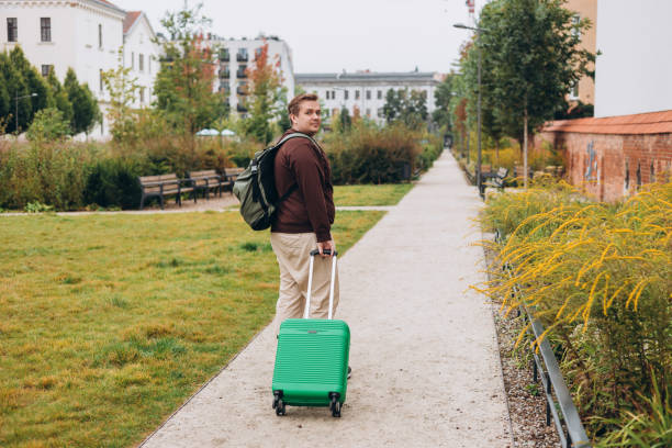 istock Happy 30s man pulling green suitcase outdoors. Rear view. Copy space. Man walking on the street with travel bag. 2178419054