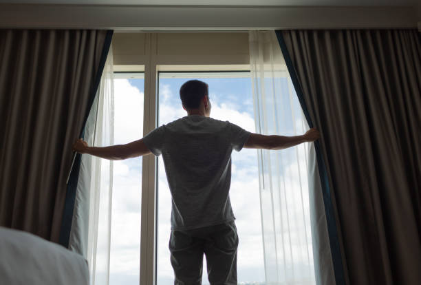 istock Happy young man opening window curtain to a sunny day. 2178444993