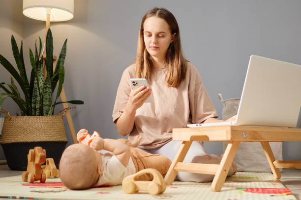 istock Busy mother using mobile phone while babysitting her daughter or son and working at home woman sitting on floor in living room engaging in online communication online browsing or internet shopping 2178450452