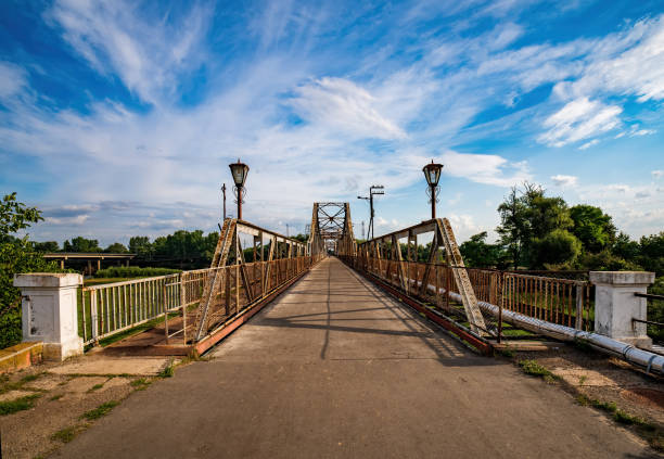 Vintage metal bridge with ornate lampposts stretches across the Dniester River in Halych, Ukraine. Dramatic sky with wispy clouds creates a picturesque scene of rural charm