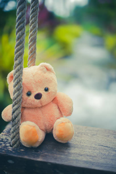 Moody low-key photograph of a teddy bear sitting alone in darkness, illuminated by subtle rim light. Minimalist composition evokes feelings of loneliness, nostalgia, childhood memories, and emotional vulnerability, with ample copy space.