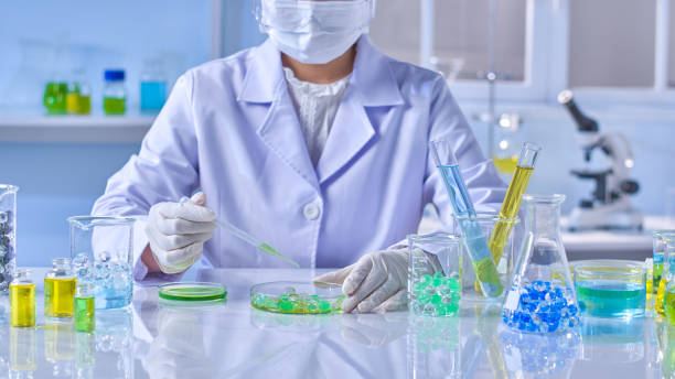 istock A female researcher with blouse coat, mask and protective eyeglass is doing an experiment in white modern lab, holding pipette to drip green essence into a glass petri dish on lab table 2178455924