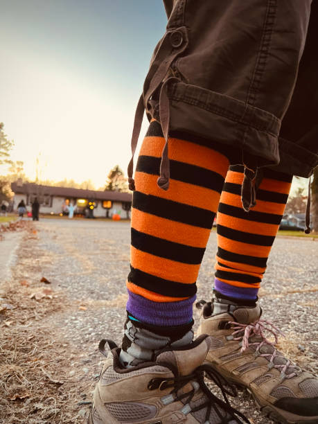 Low angle view of a women’s legs and boots only, wearing tall orange and black Halloween socks on a rural road on Halloween night during trick or treating time. Unrecognizable trick or treaters and house with Halloween decorations in the background.