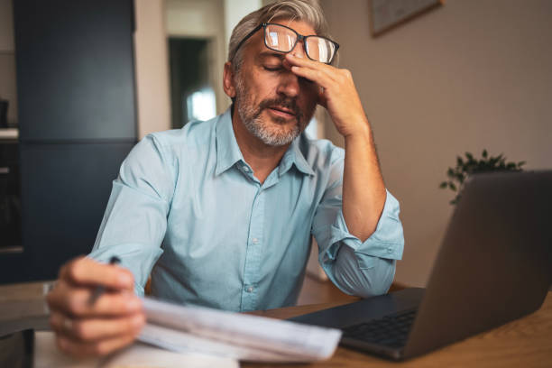 Mature man reviewing financial documents at home using laptop technology. Stressed emotion