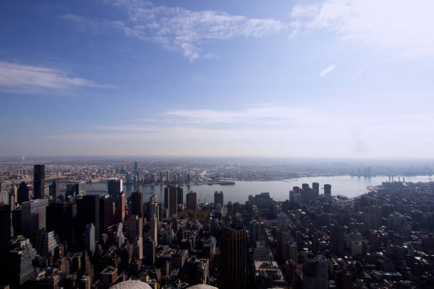 This wide-angle image offers a stunning aerial view of New York City, with towering skyscrapers in the foreground and the East River in the distance. The sprawling cityscape stretches out beneath a clear, blue sky, showcasing the dense concentration of buildings and urban life. The river adds a sense of calm and contrast to the bustling city, while bridges and distant boroughs can be seen on the horizon. This breathtaking view captures the vastness and vibrancy of the city.